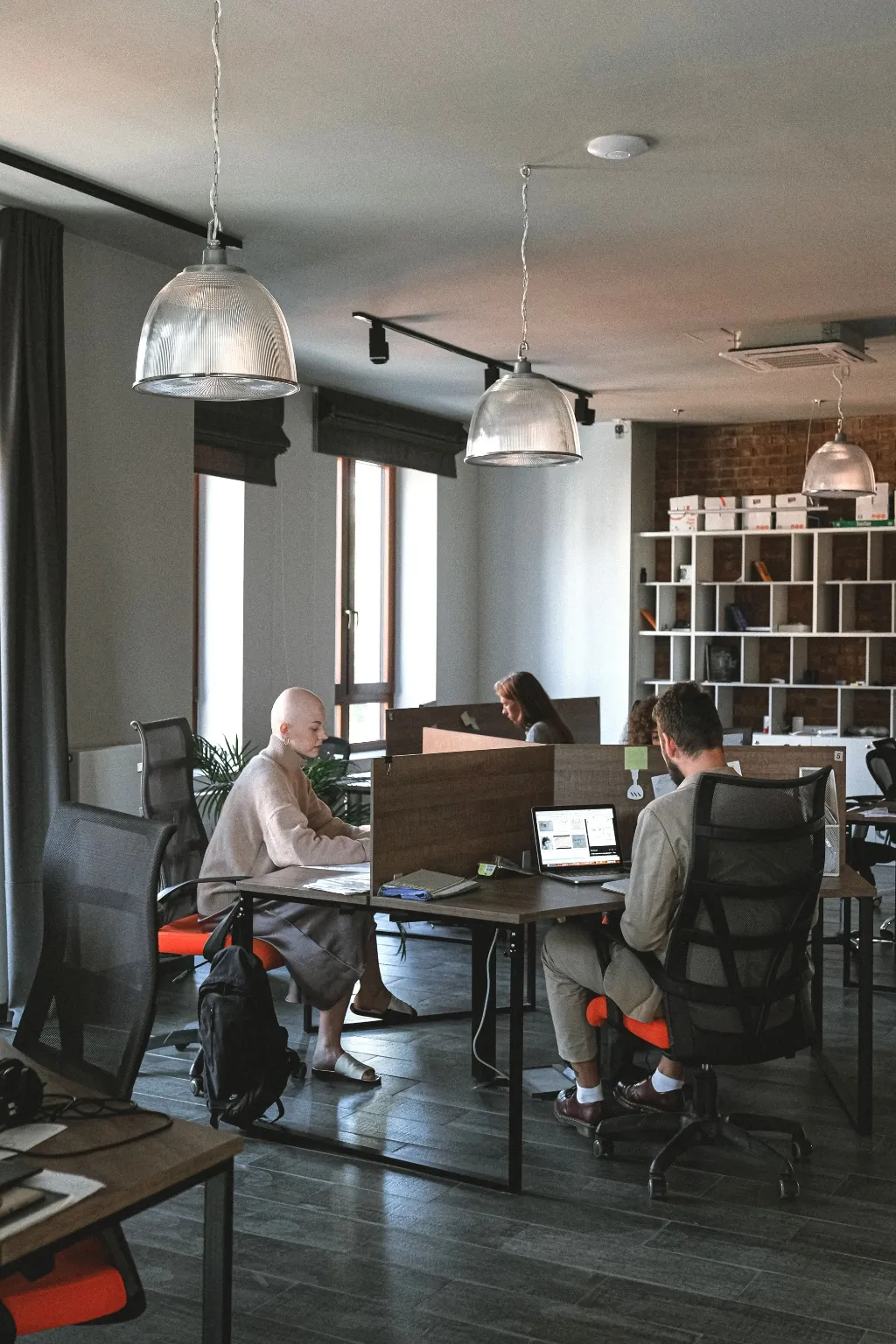 Three people work at desks in a loft-style office with pendant lights, exposed brick, and open shelving.