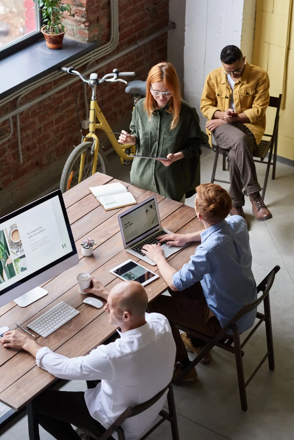Four colleagues collaborate around a wooden table with laptops, a yellow bicycle against a brick wall behind them.