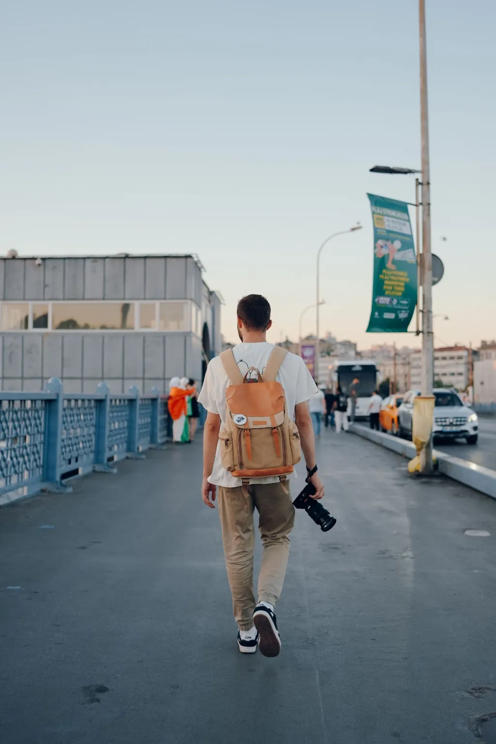 A man with a backpack and a camera walks along a city bridge at golden hour.