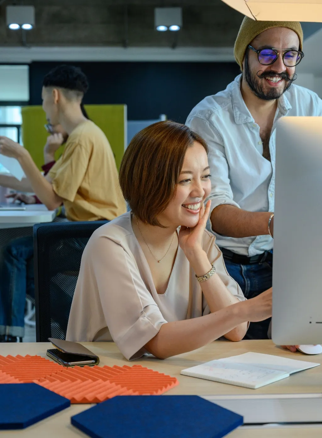 A smiling woman works at a desktop monitor in an office while a colleague stands beside her.