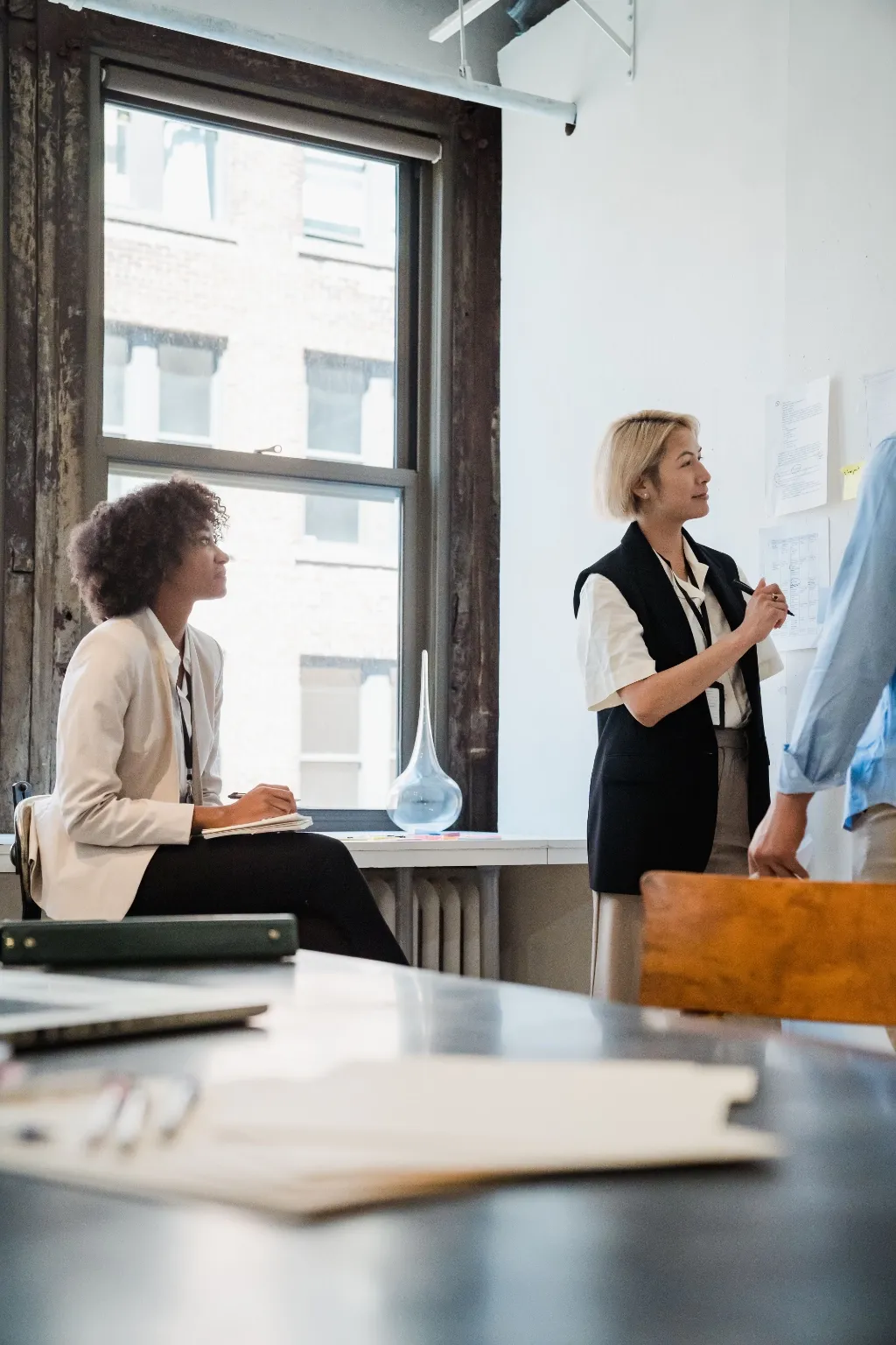 Colleagues in a meeting room, one woman presenting at a whiteboard by a window.