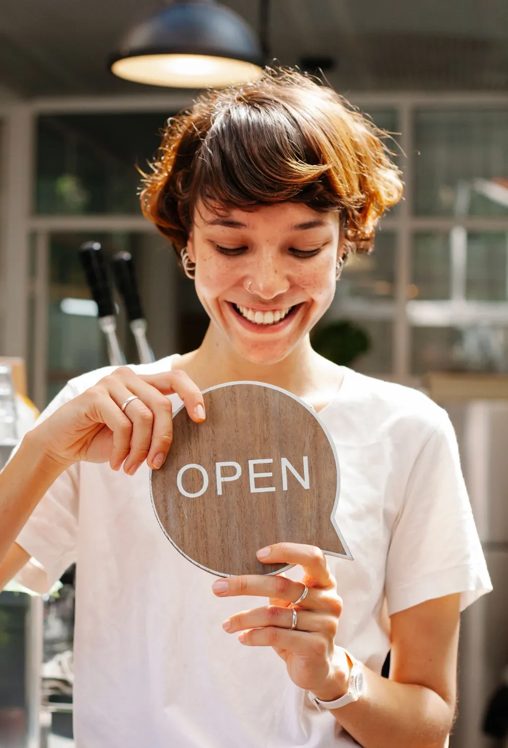 A shop owner smiles while holding up a round wooden OPEN sign in a bright storefront.
