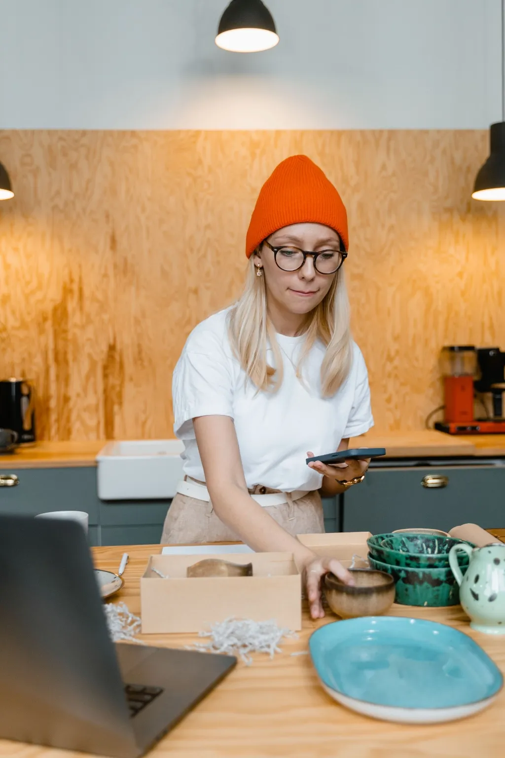 A maker checks an order on her phone while boxing handmade ceramics at a studio bench.