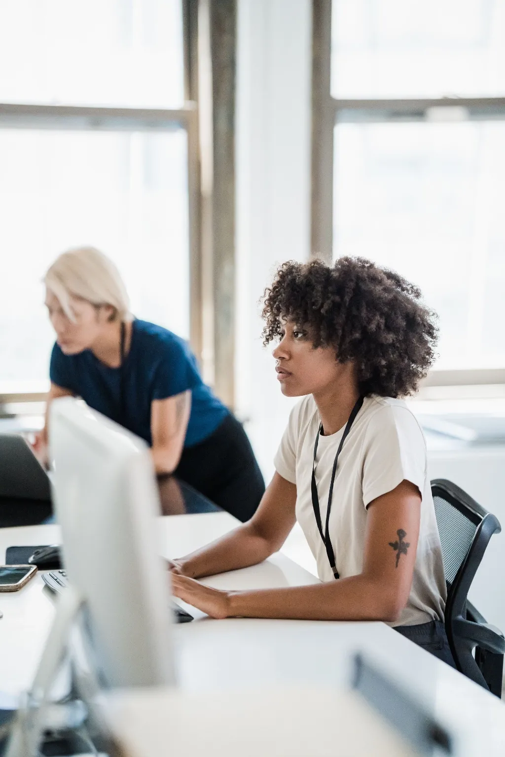 A woman with curly hair works at a desktop computer in a bright office while a colleague works at a desk behind her.