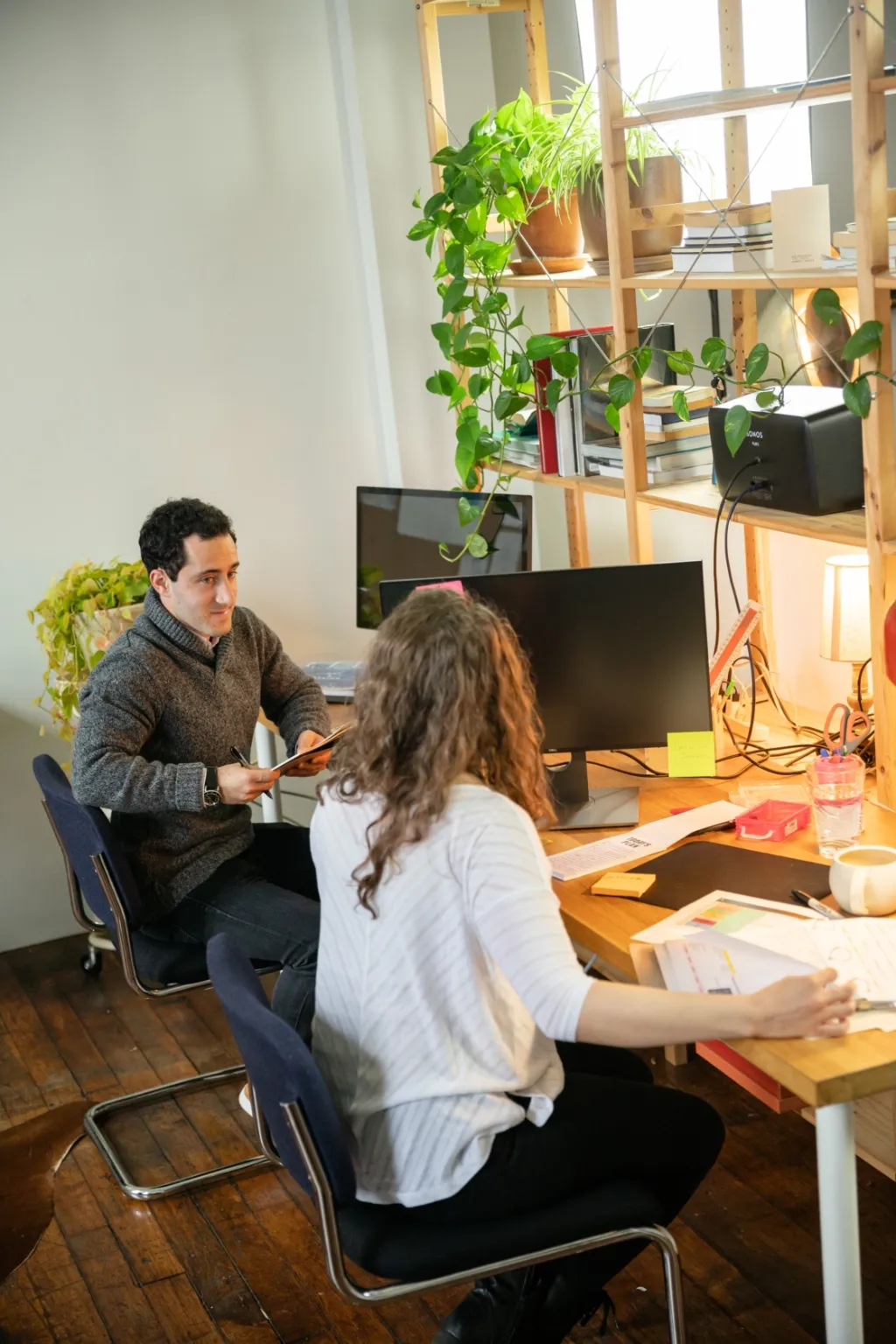 A man and a woman work together at a desk in a plant-filled studio, the man passing something across the table.