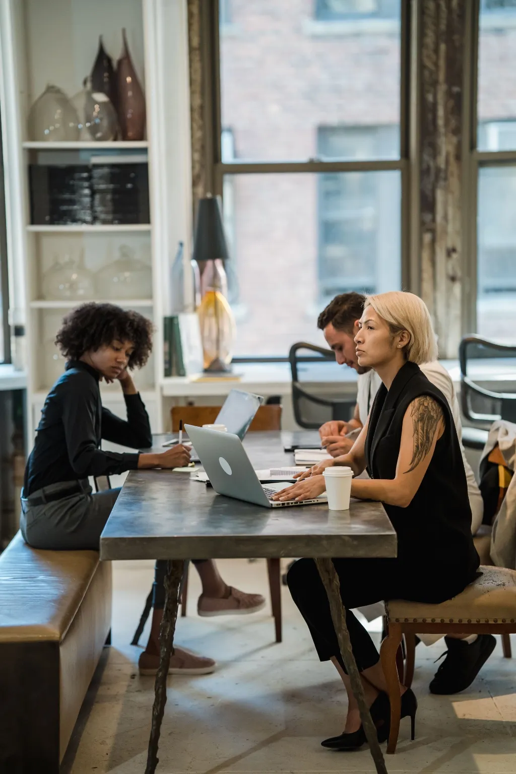 Three colleagues work around a long table with a laptop and coffee in a loft-style office.