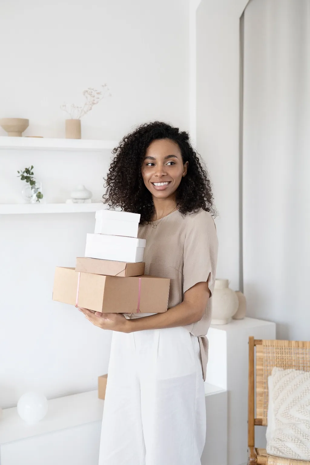 A smiling woman holds a stack of parcels in a bright, minimal room.