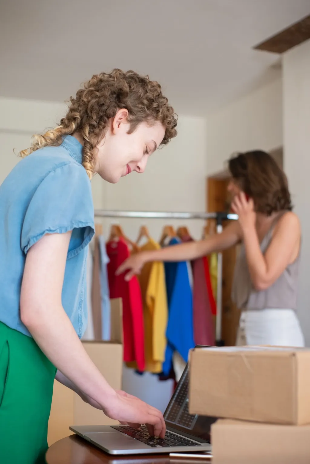 A boutique owner packs an online order on a laptop while a customer browses a rack of clothes behind her.